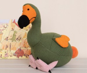 Knitted moss green dodo soft toy with painting of dinosaurs behind